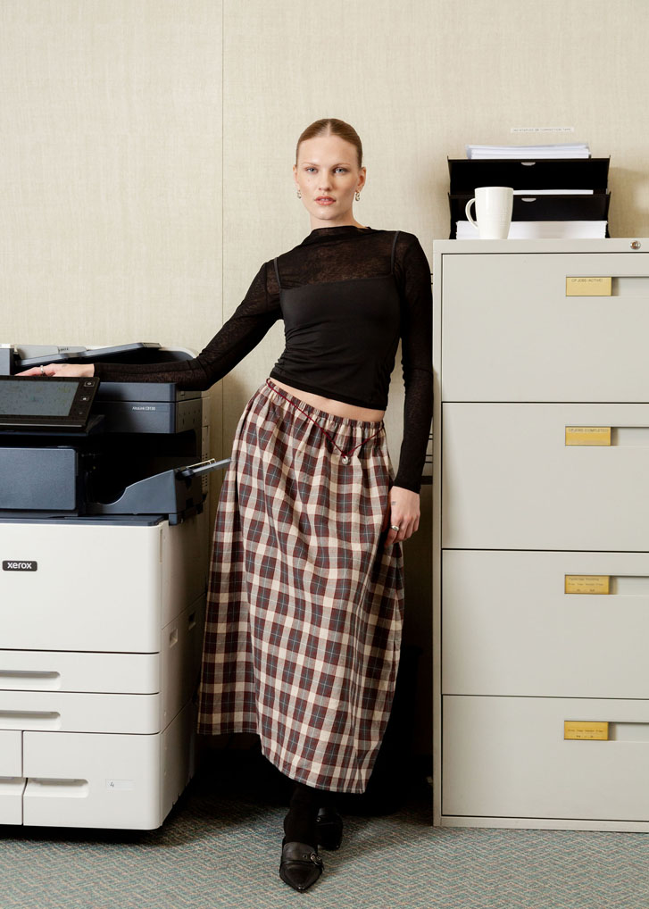 lady fashionably standing in an office wearing a long plaid skit and black see-through long sleeve with a black tank camisol