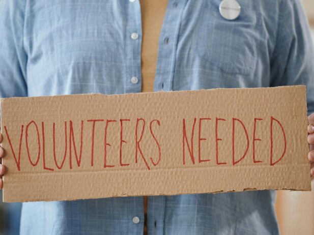 a woman in a jean jacket holding up a box sign that reads "Volunteers Needed"