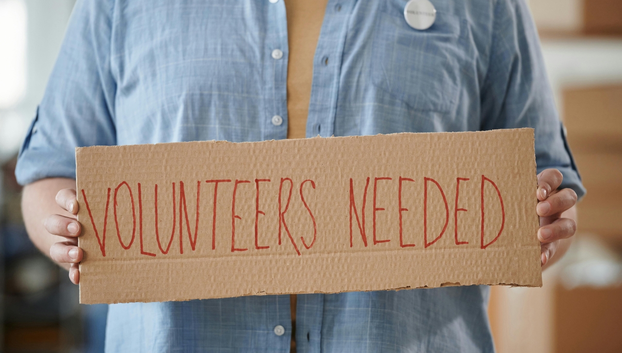 a woman in a jean jacket holding up a box sign that reads "Volunteers Needed"