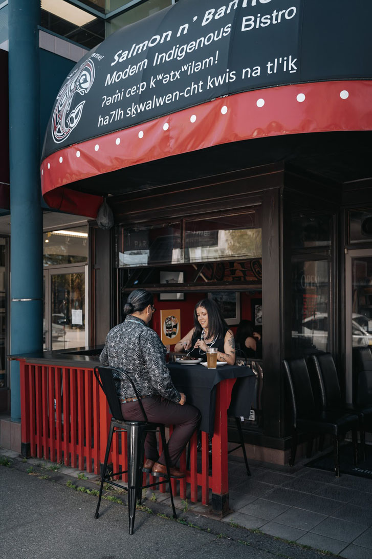 a man and woman seated and eating a cuisine at an outside table over a restaurant banner that says: 'Salmon N’ Bannock Modern Indigenous Bistro'