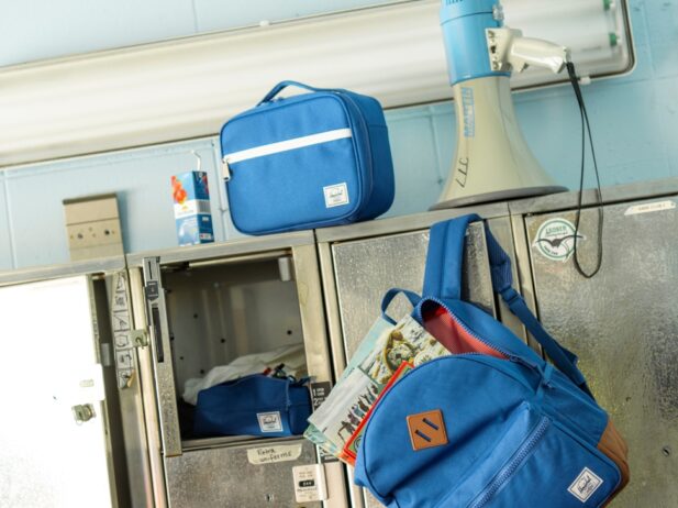 a steel school locker stand with an open locker next to a hanging open blue back-to-school bag, a blue and white megaphone, a blue lunch bag and a box juice on top of the locker stand