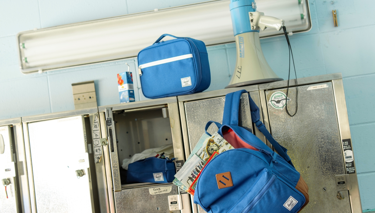 a steel school locker stand with an open locker next to a hanging open blue back-to-school bag, a blue and white megaphone, a blue lunch bag and a box juice on top of the locker stand