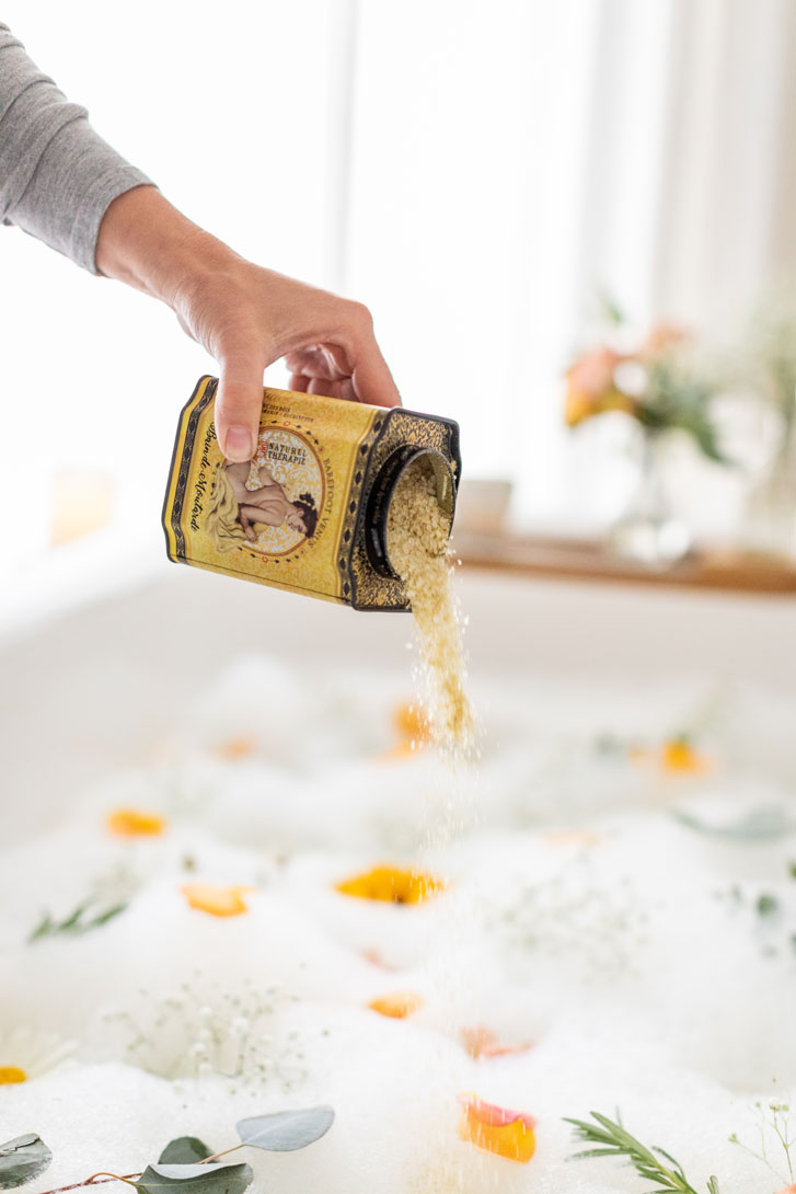 woman hand poring out the contents of a mustard bath spa product into a flowered bubbling bath tub