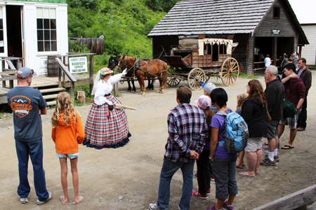 people gathered around a woman dressed in a traditional gown with horses and a carriage behind her at a historical sites