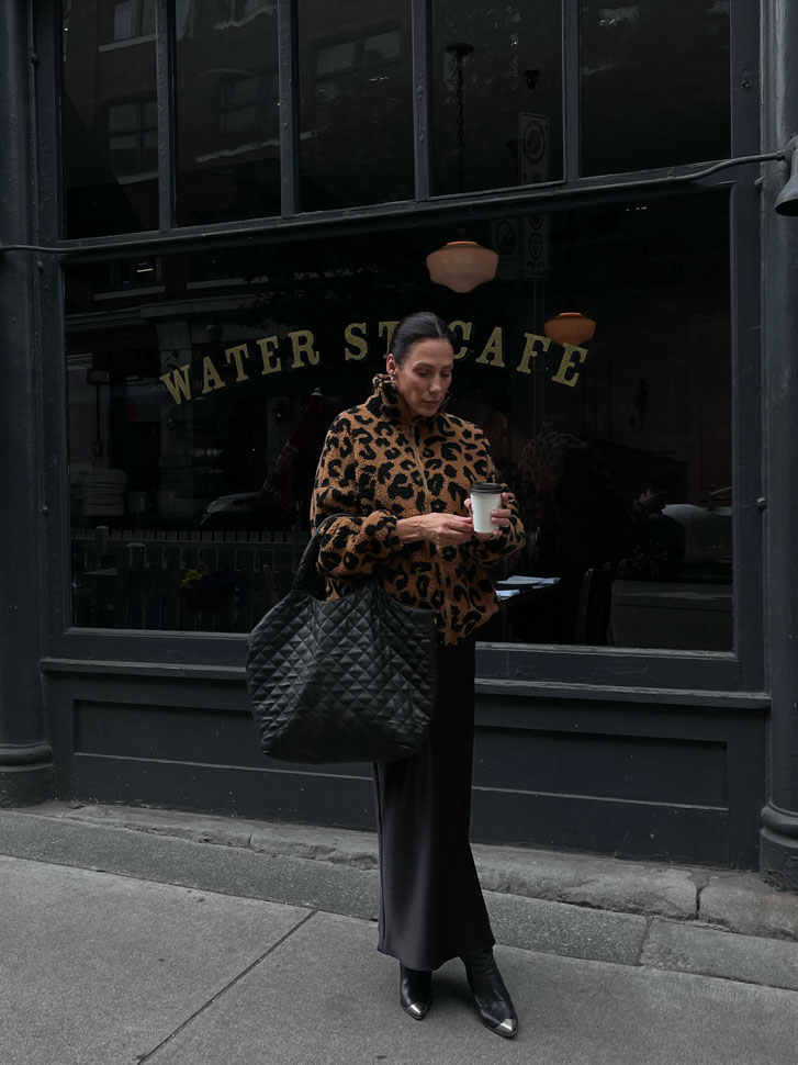 woman in luxury fashion leopard patterned brown coat and a long black silk skirt with a black bag and black boats while standing in front of a cafe