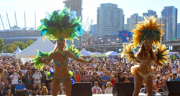 2 dancing ladies wearing colorful animal feathers with a huge crowd behind