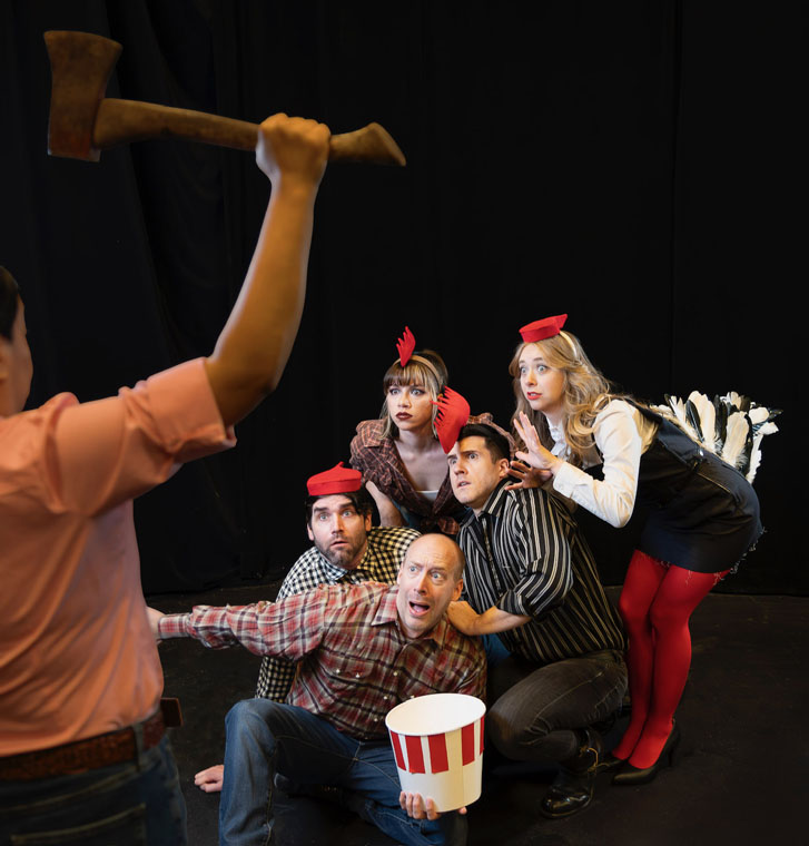 a group of theatre performers with rooster hats seated on a stage with scary faces while a man stands in front with an axe