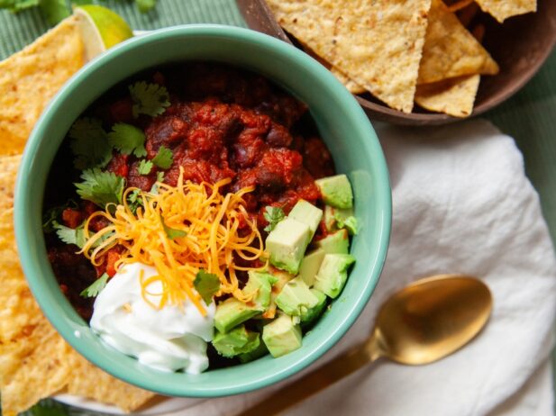 a bowl of chili with a topping of sliced avocado, sliced carrots, cilantro & sour cream and a bowl of tortilla chips