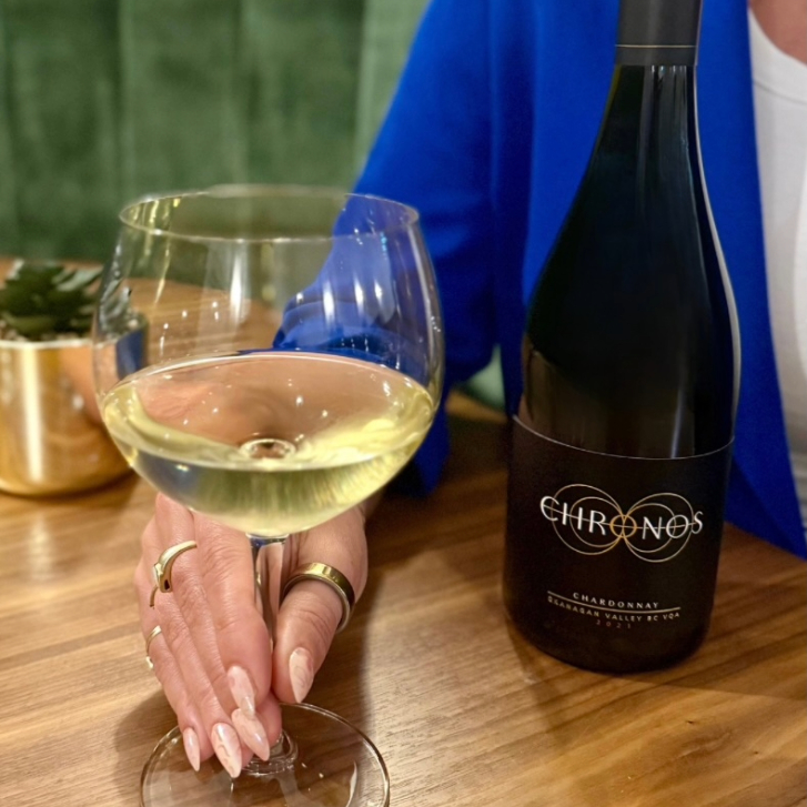 person wearing a blue blazer holding the stem of a wine glass with white wine in it with a Chronos bottle behind