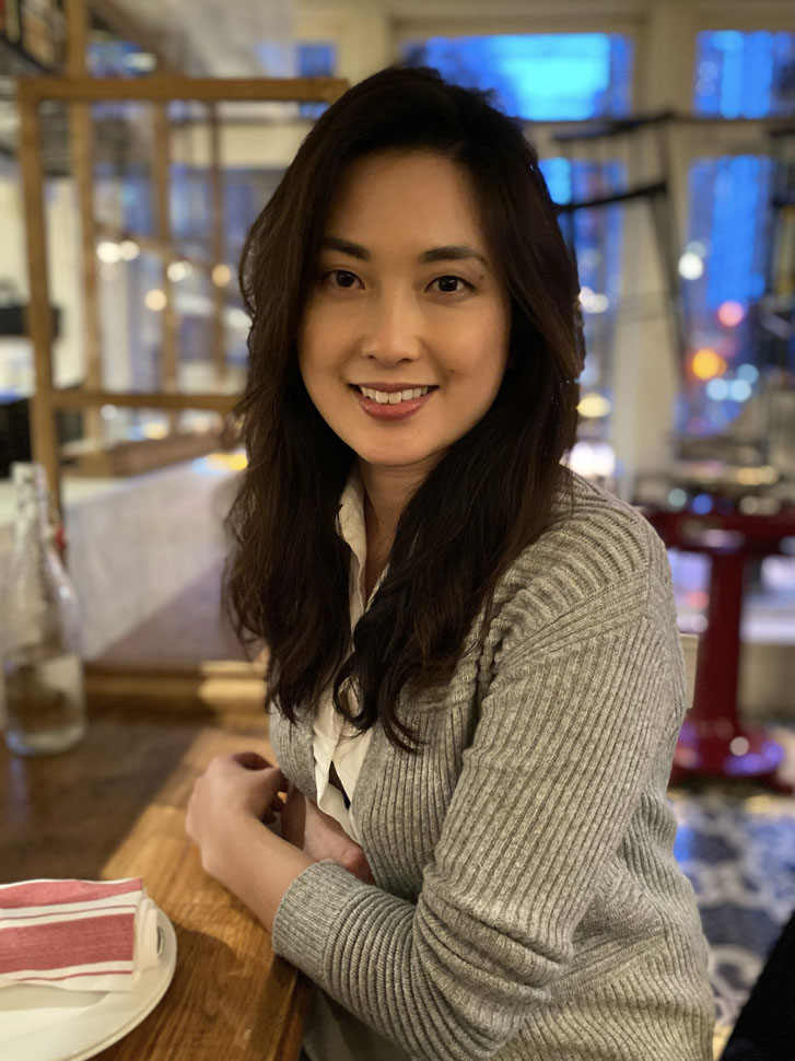 a forward-smiling asian woman sitting inside a food startup restaurant with a folded dish towel on her plate