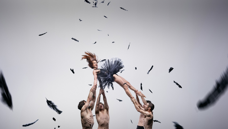 a theatre dancer in ballet dress thrown up by three shirtless dancer guys with black confetti in the air