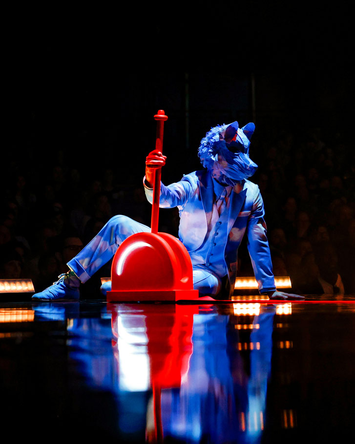 man in a dog mask seated on a dark stage with his hands on a large red engine stopper 