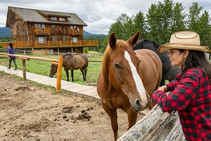 woman in cowboy hat standing beside horses in a stable grazing and a big wooden building lodge at one end