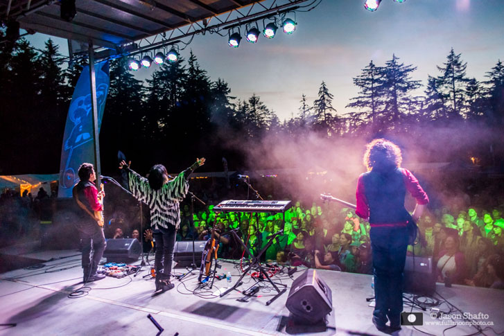 two performers playing guitars and a woman performer in the middle with arms wide open at an outdoor stage event