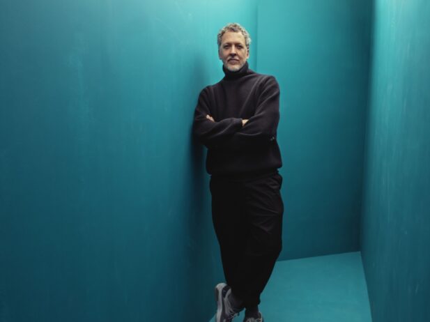 a standing man with his left leg crossed over the other, leaning on a blue wall inside a lit blue room