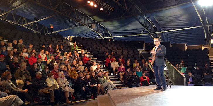 a man on a stage speaking to a room filled with people seated