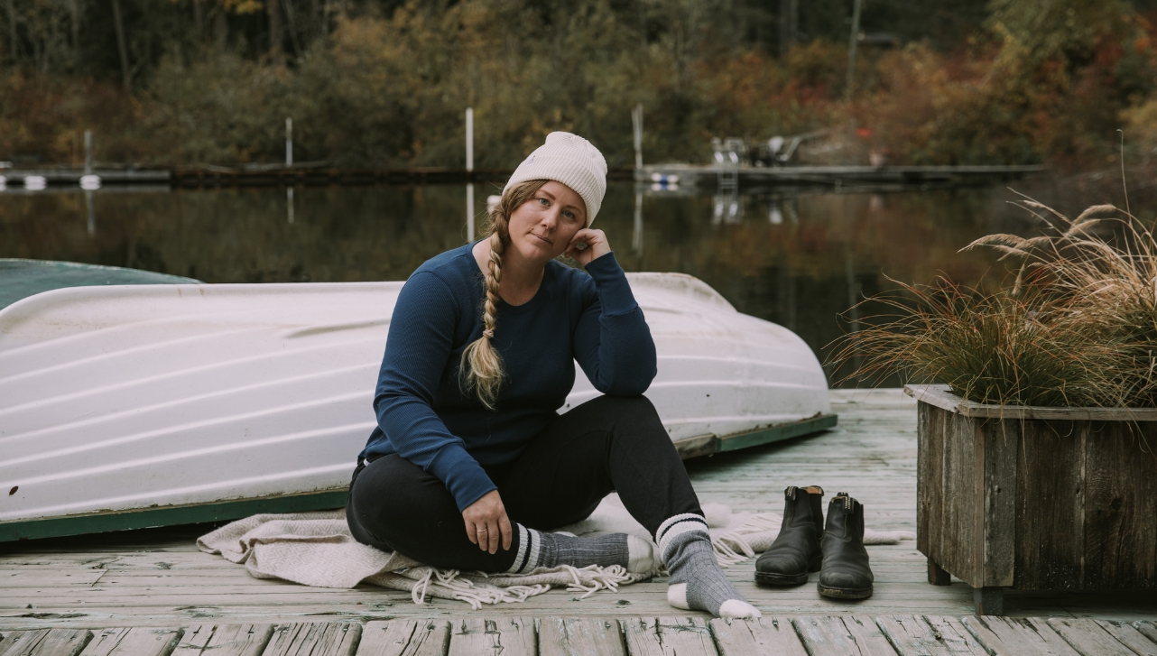 woman in covered fashion clothing, beanie, and socks, sitting on a deck with her black boots beside her and a turned-over boat behind her