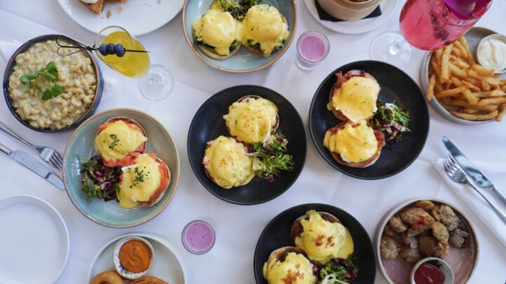 flatlay view of eggs benedict benny dishes, a macaroni food dish, fries, onion rings and potato dishes all on different plates on a white cloth table