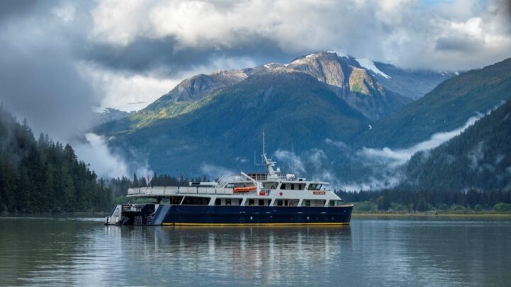 a ship sailing on an ocean with high mountains and clouds behind it with the Great Bear Rainforest