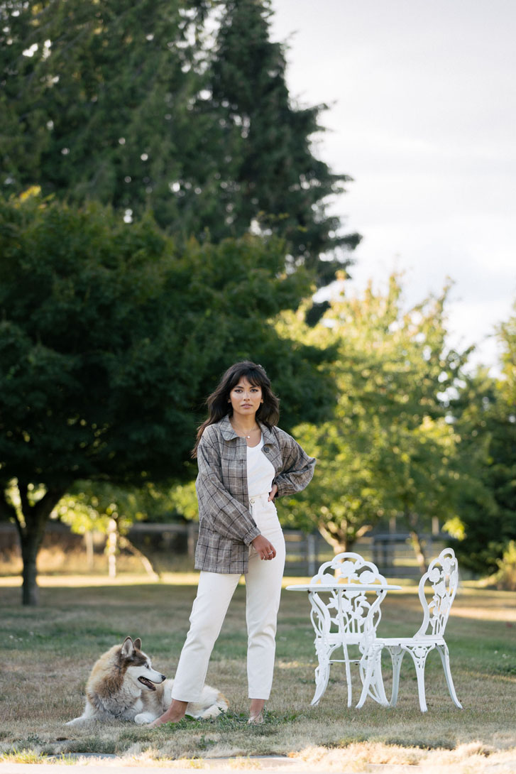 woman in white top, cream jean and neutral shade jacket, standing beside her dog on a leveled grass