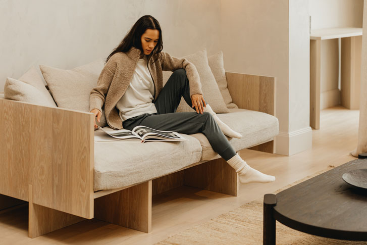 lady in a clothing brand sweaters and sweat pants while seated on a couch looking through a magazine