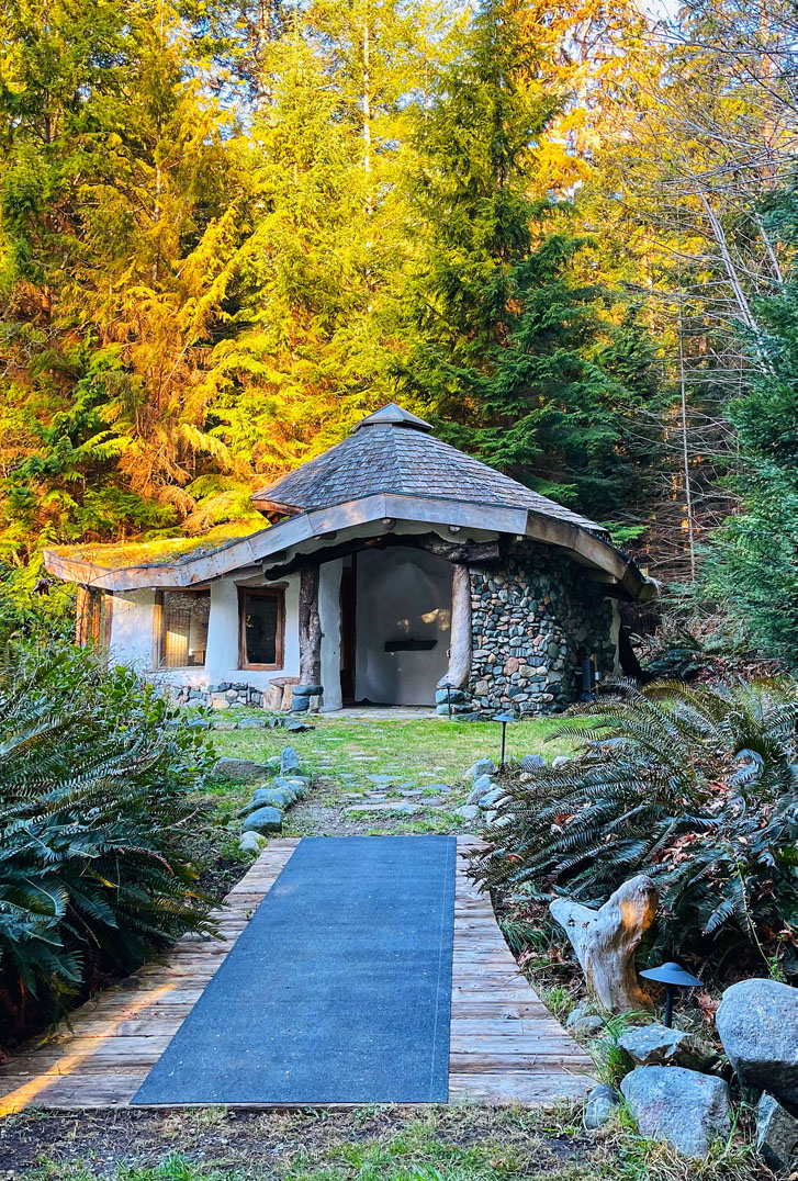 a small stone-and-wood-styled building with tall trees behind it and a walkway in front with flowers on each side