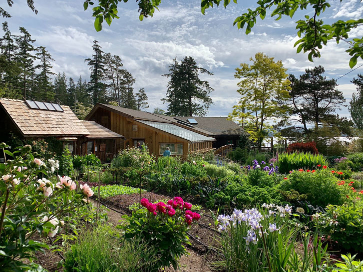 a woman and man standing in front of a 2-set lodge building with a green colorful flower garden in front and trees and ocean behind