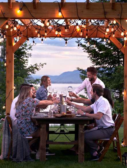 a group of guys and ladies clinking their glasses of wine while seated round a wooden table overlooking a river