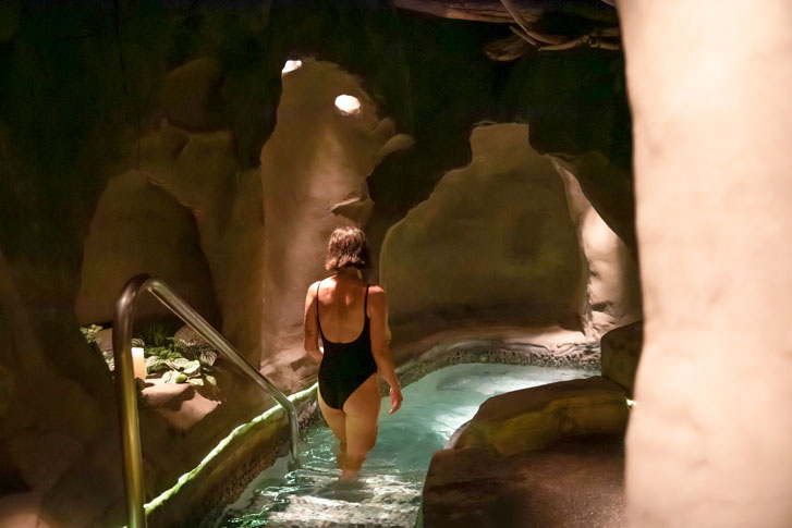 woman in a black swimsuit walking down the water stairway into a narrow pool in a clay-like building with rays of sunlight coming through the tiny carved holes in the building