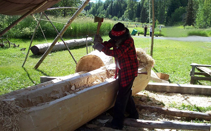 a wood worker carving a boat-like structure with an axe 