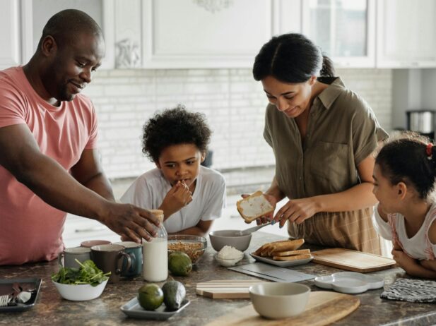 father, mother and two kids at the kitchen island counter with bread slices, avocados, cups, vegetables, and a jar of milk on the counter