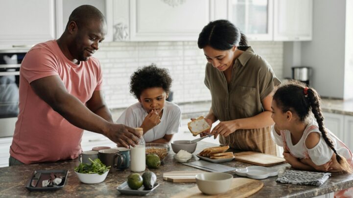 father, mother and two kids at the kitchen island counter with bread slices, avocados, cups, vegetables, and a jar of milk on the counter