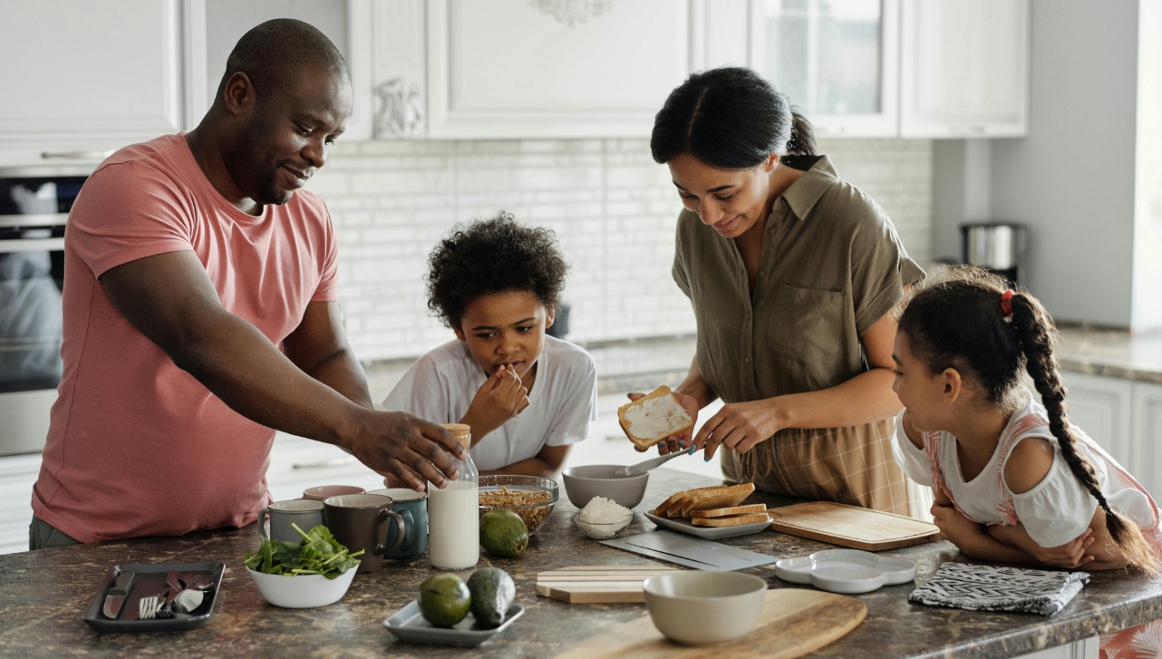 father, mother and two kids at the kitchen island counter with bread slices, avocados, cups, vegetables, and a jar of milk on the counter