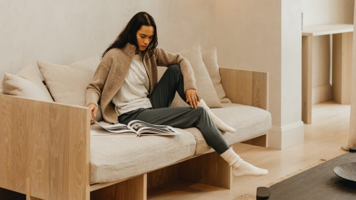 lady in a clothing brand sweaters and sweat pants while seated on a couch looking through a magazine