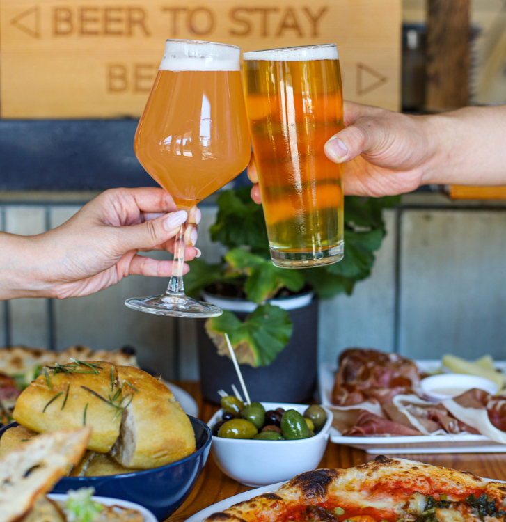 two people cheersing with glasses of beer with a table full of food at Luppolo in Vancouver
