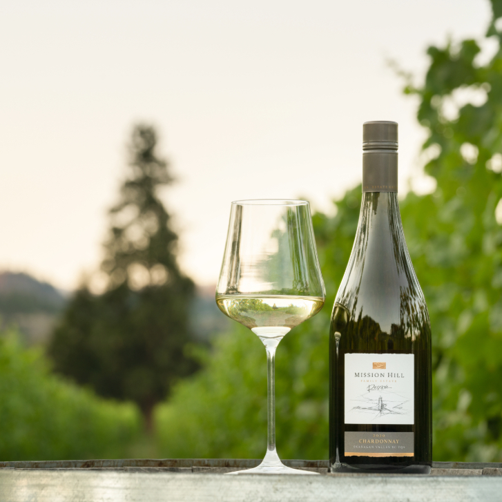 glass of chardonnay next to a bottle on a table outside with trees in the background