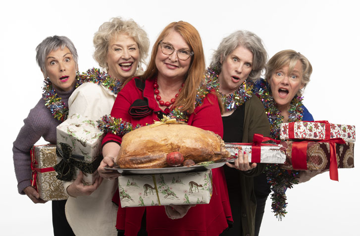 five elderly women holding wrapped Christmas gift boxes while the woman in the middle also holds a tray of huge turkey