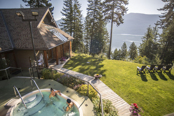 two women talking in a pool by a resort lodge building on a hill with sparse trees, overlooking the mountains and a lake