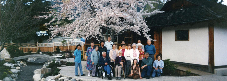 a group of elderly Japanese Canadians standing and sitting together under a tree at a historical sites