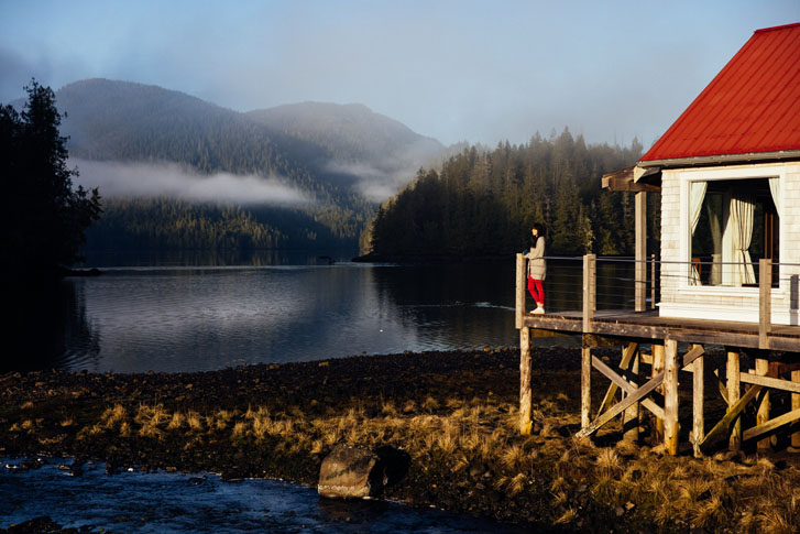 woman standing on the deck of an elevated wooden house on one end of a wide rainforest by a huge lake