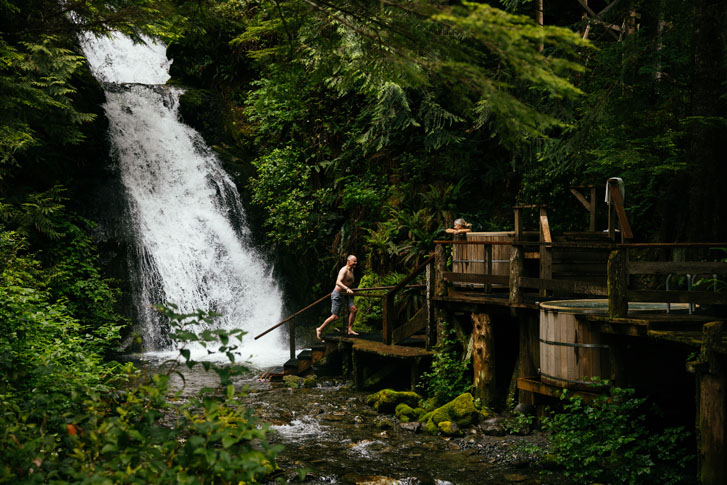 man walking up a wooden stairway from a huge waterfall to meet a woman in a wooden round bathtub pool in the rainforest