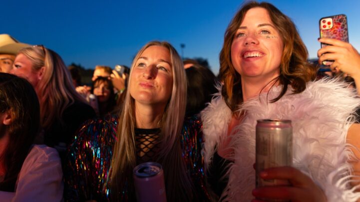 two ladies holding drink cans while watching an event alongside a group of other people outdoor