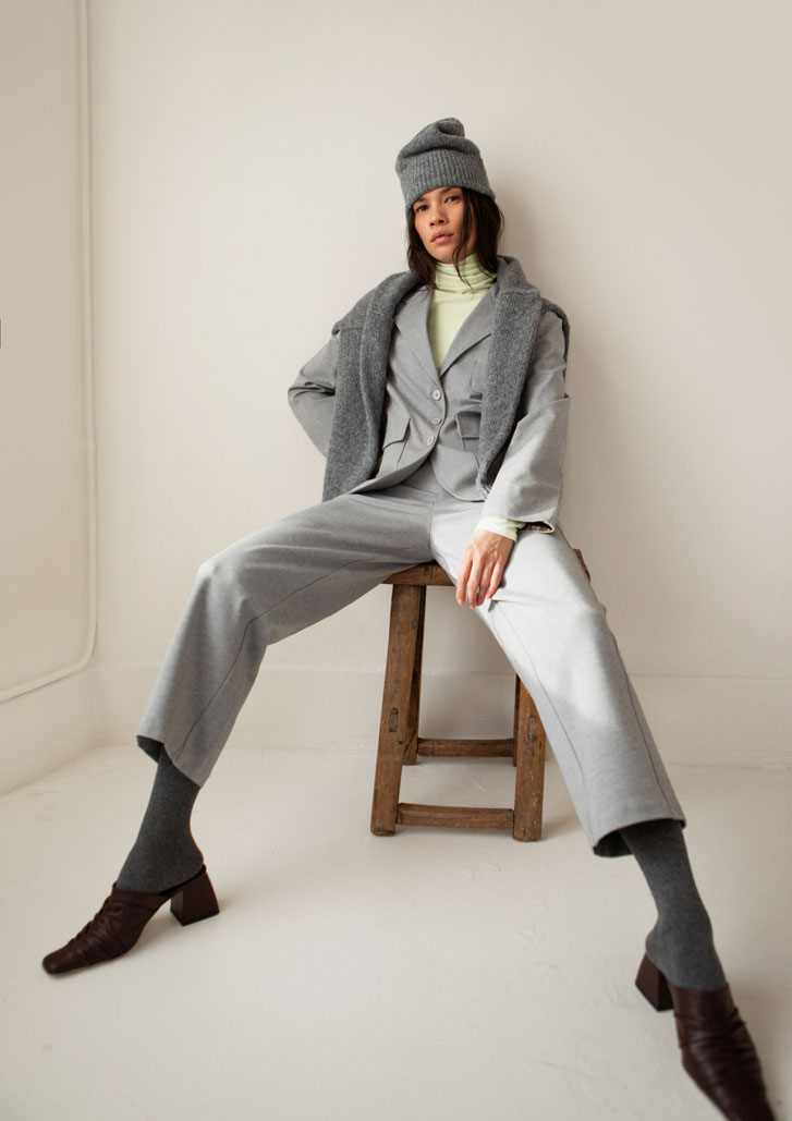 woman sitting on a chair fahionably and wearing corporate staple clothing with a grey beanie and a cardigan over her shoulders