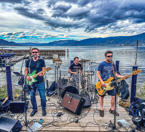 a band of guitarist and a drummer performing on a wooden deck behind a huge lake