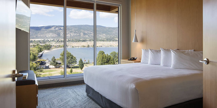 doors opening into white bed in a bedroom with tall transparent glass windows and a flat scrren hanging on the wall in front of the bed