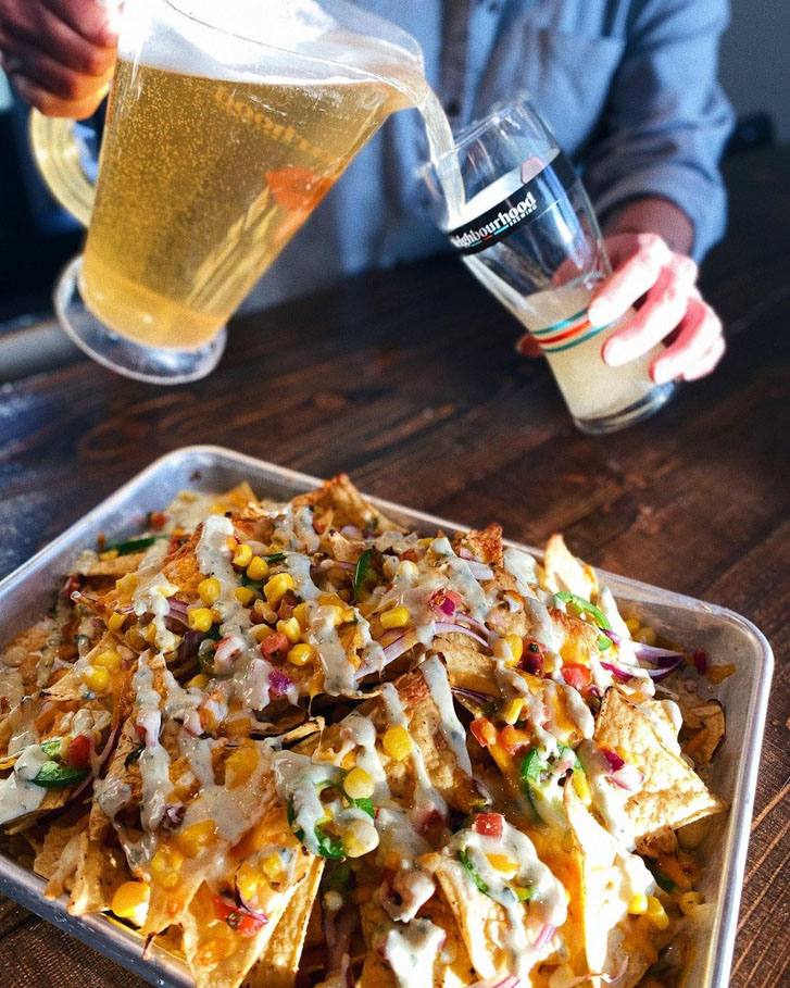 man pouting a beer from a jug into a glass with a tray of mixed veggie-dressed nachos in front of him