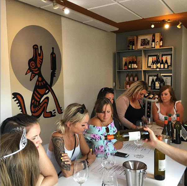 a group of ladies seated around a table with a hand pouring a bottle of wine into empty glasses on the table