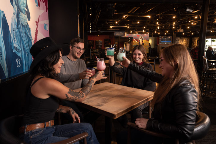 a group of girls and a guy toasting around a table with glasses of colorful drinks