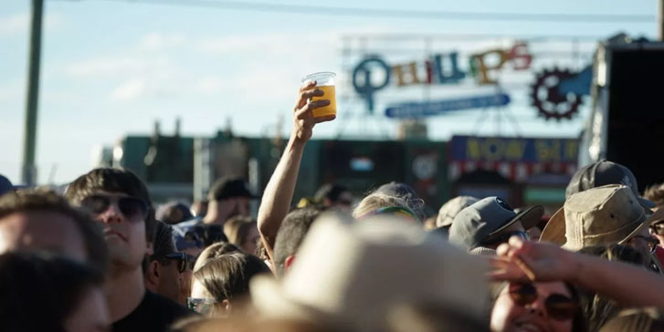 a crowd in the sun and an outstretched arm holding a glass of juice above the heads in the crowd towards the sky