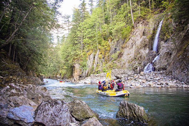 five paddlers in live jackets in a colorful raft holding up their paddles together in a high five on a moving lake with rocks and cliffs on both sides of the lake